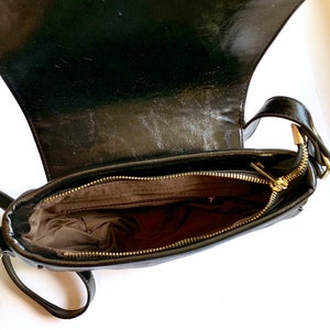 Puede incluir: Bolso bandolera de cuero negro con cremallera dorada y cierre de solapa. El bolso est&aacute; abierto y el interior est&aacute; forrado con tela marr&oacute;n.