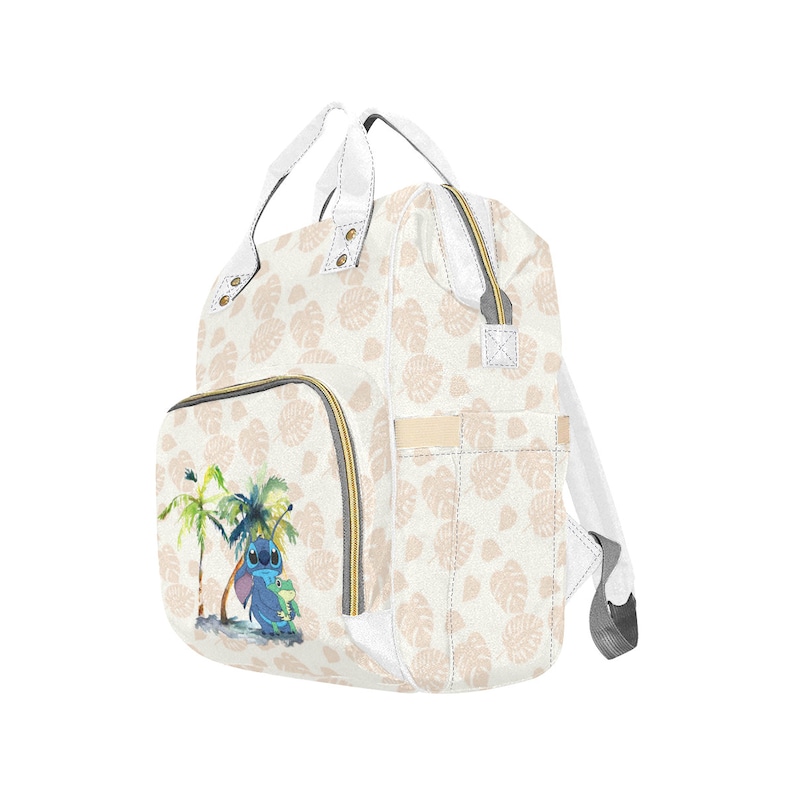 Puede incluir: Una bolsa de pa&ntilde;ales blanca con un estampado floral marr&oacute;n claro y un personaje azul Stitch de Lilo & Stitch en la parte delantera. La bolsa tiene un bolsillo con cremallera y un asa superior.
