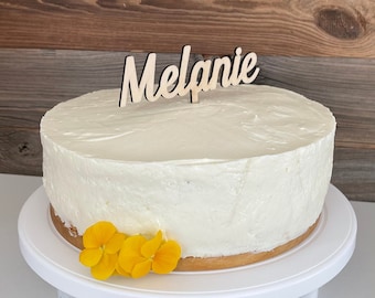May include: A white cake with a wooden topper that says "Melanie" and yellow flowers on a white cake stand.