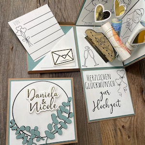Op de afbeelding: Een witte kaart met een groene krans en de tekst "Daniela & Nicole" in gouden letters. Een tweede kaart heeft de tekst "Herzlichen Gl&uuml;ckwunsch zur Hochzeit" in zwarte letters.