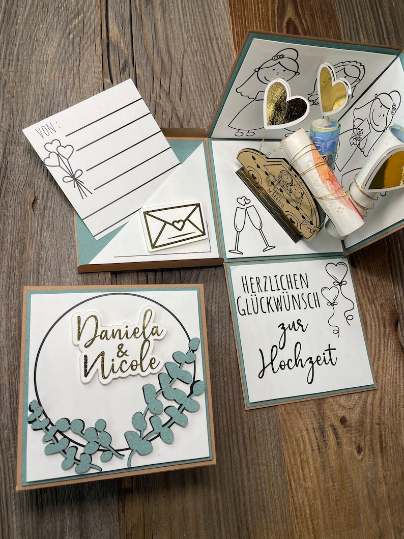 Op de afbeelding: Een witte kaart met een groene krans en de tekst "Daniela & Nicole" in gouden letters. Een tweede kaart heeft de tekst "Herzlichen Gl&uuml;ckwunsch zur Hochzeit" in zwarte letters.