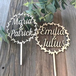 May include: Two wooden cake toppers with leafy wreaths and the names "Maria & Patrick" and "Emilia & Julius" written in cursive.