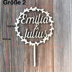 May include: A wooden cake topper with a wreath design and the names "Emilia & Julius" in a cursive font. The topper is 20.5 inches tall and 13.5 inches wide.