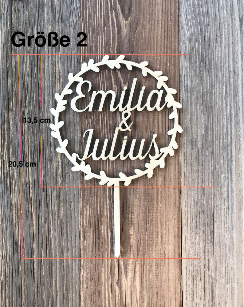 May include: A wooden cake topper with a wreath design and the names "Emilia & Julius" in a cursive font. The topper is 20.5 inches tall and 13.5 inches wide.