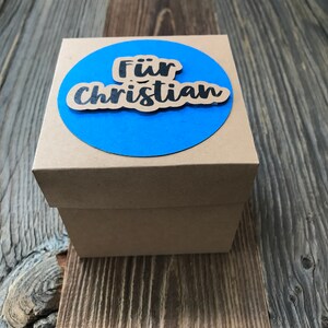 May include: A brown cardboard gift box with a blue circle on top. The circle has the words "F&uuml;r Christian" written in black.