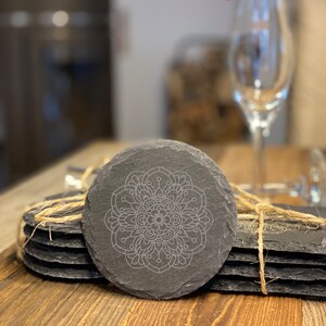 May include: Set of six black slate coasters with a white mandala design. The coasters are tied together with twine and the text "Mandala 1" is printed on a wooden surface.