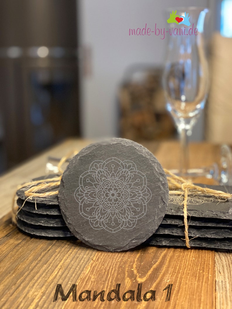 May include: Set of six black slate coasters with a white mandala design. The coasters are tied together with twine and the text "Mandala 1" is printed on a wooden surface.