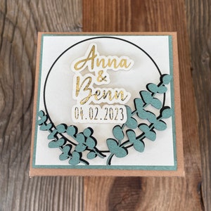 Op de afbeelding: Een witte kaart met een gouden cirkel en groene bladeren eromheen. De tekst "Anna & Benn 04.02.2023" is geschreven in gouden letters.