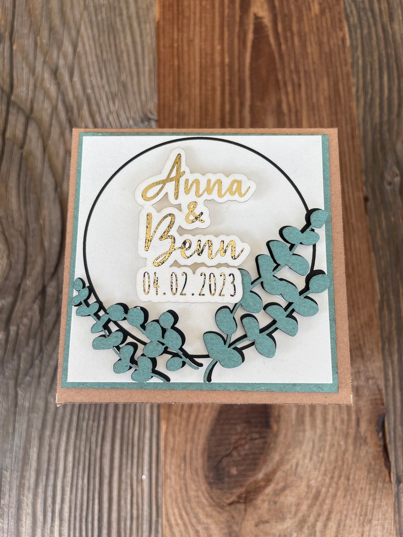 Op de afbeelding: Een witte kaart met een gouden cirkel en groene bladeren eromheen. De tekst "Anna & Benn 04.02.2023" is geschreven in gouden letters.