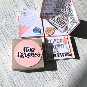 May include: A handmade birthday card with multiple layers and pop-up elements. The card features a pink and white color scheme, with handwritten text and decorative stickers. The card is personalized with the name "Lorena" and includes a small gift box.