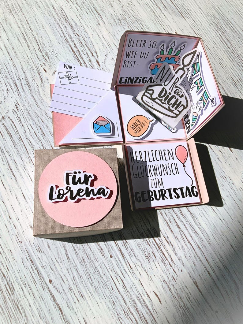 May include: A handmade birthday card with multiple layers and pop-up elements. The card features a pink and white color scheme, with handwritten text and decorative stickers. The card is personalized with the name "Lorena" and includes a small gift box.
