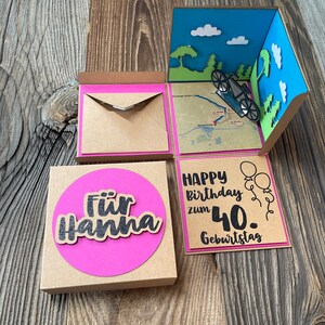 May include: A brown cardboard box with a pink circle on the front that says "F&uuml;r Hanna". The box is open and inside is a pop-up card with a blue sky, white clouds, green trees, and a bicycle. The card also has a pink border and says "Happy birthday zum 40. Geburtstag".