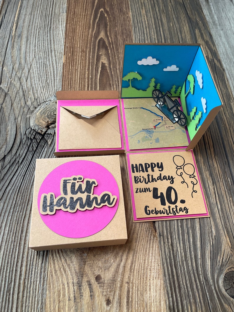 May include: A brown cardboard box with a pink circle on the front that says "F&uuml;r Hanna". The box is open and inside is a pop-up card with a blue sky, white clouds, green trees, and a bicycle. The card also has a pink border and says "Happy birthday zum 40. Geburtstag".