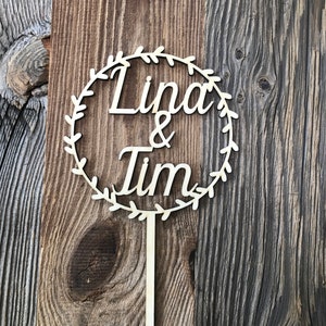 May include: A wooden cake topper with a circle design and the names "Linda & Tim" written in cursive. The circle is decorated with a leafy wreath.
