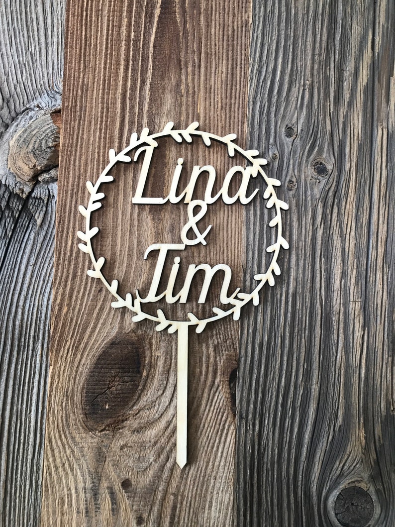 May include: A wooden cake topper with a circle design and the names "Linda & Tim" written in cursive. The circle is decorated with a leafy wreath.