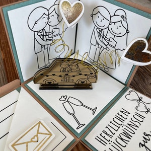 Op de afbeelding: Een teal en bruine explosiebox met een pop-up auto waarop "Just Married" staat en een stel erin. De doos is versierd met verschillende andere decoraties, waaronder harten, champagne glazen en een envelop.