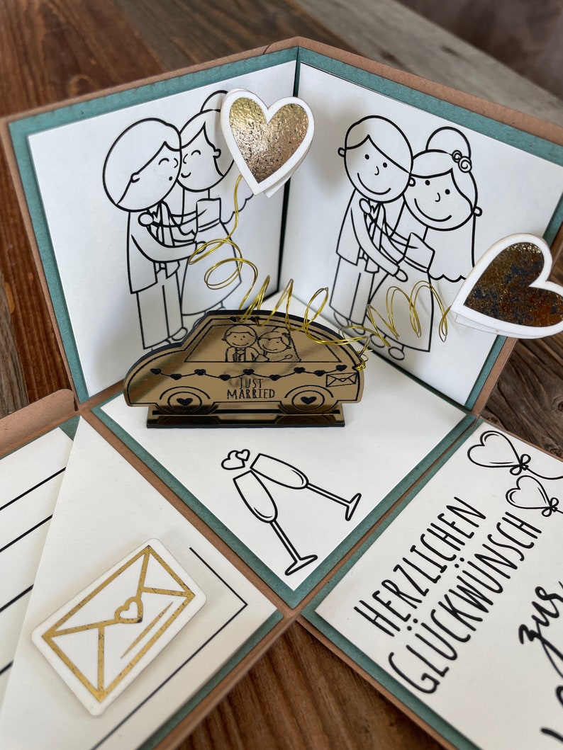 Op de afbeelding: Een teal en bruine explosiebox met een pop-up auto waarop "Just Married" staat en een stel erin. De doos is versierd met verschillende andere decoraties, waaronder harten, champagne glazen en een envelop.