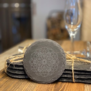 May include: A set of six black slate coasters with a white mandala design. The coasters are stacked and tied together with twine. The text "Mandala 4" is printed on the wooden surface below the coasters.
