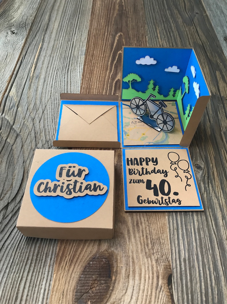 May include: A brown cardboard box with a blue circle that says "F&uuml;r Christian" on the front. The box is open and reveals a pop-up card with a bicycle, trees, and clouds. The card also has the text "Happy Birthday zum 40. Geburtstag" on it.