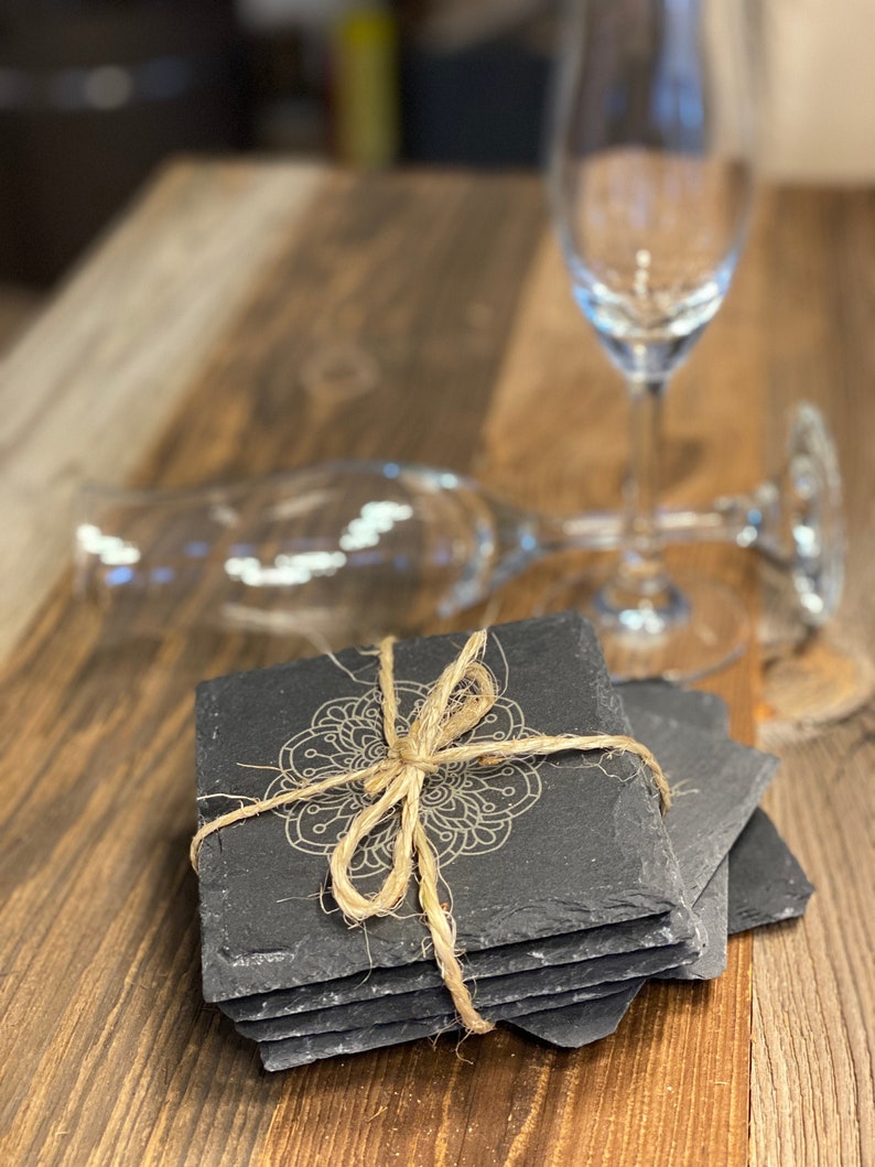 May include: A set of six black slate coasters with a decorative mandala design. The coasters are tied together with twine.