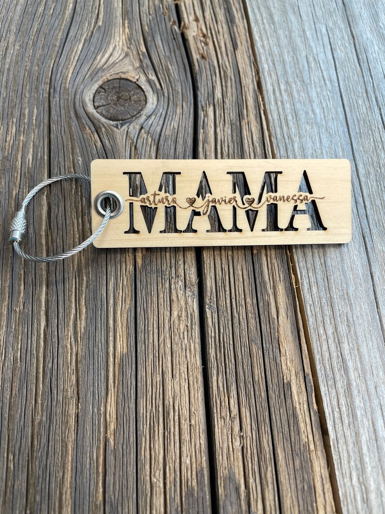 May include: Wooden keychain with the word "MAMA" engraved on it. The keychain has a silver key ring and the names "Arturo", "Javier", and "Vanessa" are engraved below the word "MAMA" with a heart symbol between each name.