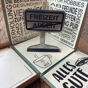 Puede incluir: Una tarjeta marr&oacute;n y blanca con el texto "Die besten W&uuml;nsche" y "Zeit f&uuml;r Hobbies" en ella. La tarjeta est&aacute; doblada por la mitad y tiene un letrero de metal que dice "Freizeit Arbeit". El letrero est&aacute; sobre un peque&ntilde;o soporte de metal. Hay una taza peque&ntilde;a con el texto "Zeit f&uuml;r mich" en ella.