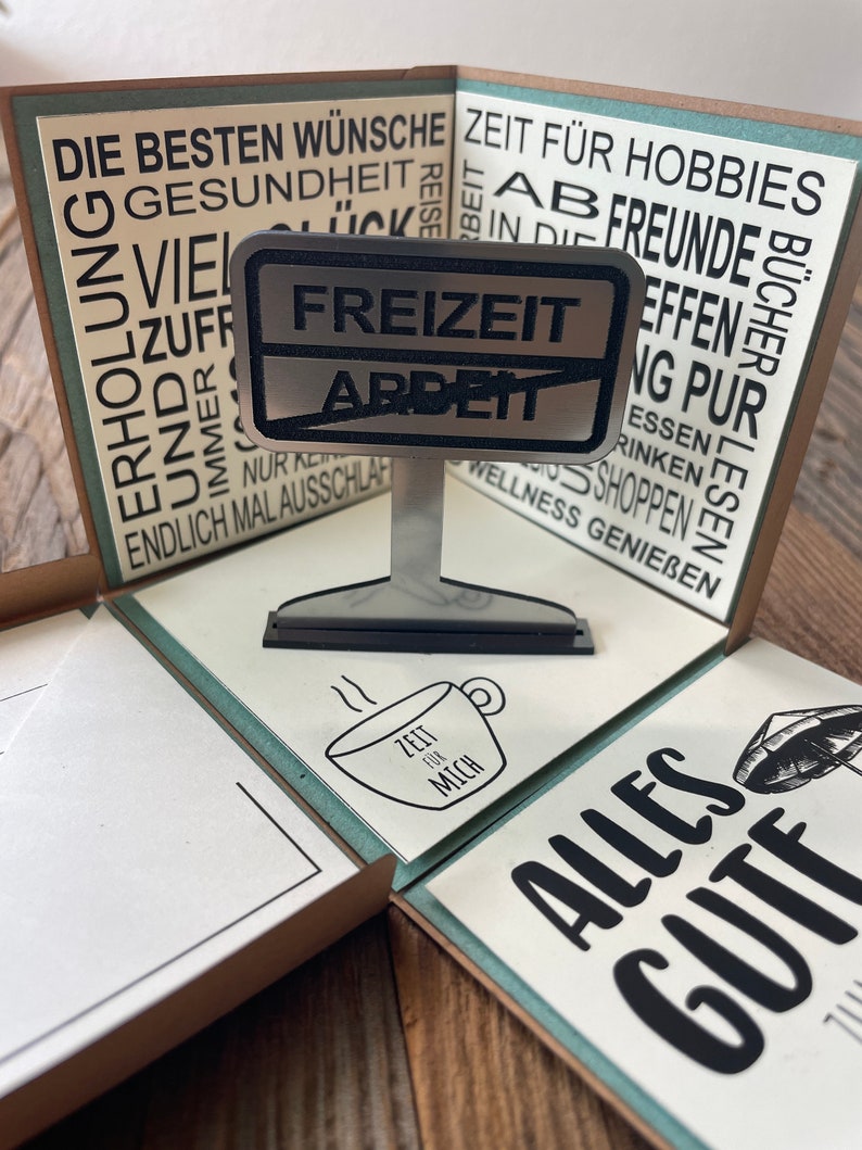 Puede incluir: Una tarjeta marr&oacute;n y blanca con el texto "Die besten W&uuml;nsche" y "Zeit f&uuml;r Hobbies" en ella. La tarjeta est&aacute; doblada por la mitad y tiene un letrero de metal que dice "Freizeit Arbeit". El letrero est&aacute; sobre un peque&ntilde;o soporte de metal. Hay una taza peque&ntilde;a con el texto "Zeit f&uuml;r mich" en ella.