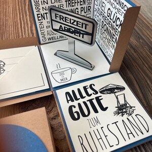 Puede incluir: Una tarjeta blanca con texto negro que dice "Alles Gute zum Ruhestand" con una ilustraci&oacute;n en blanco y negro de una sombrilla de playa y una silla de playa. La tarjeta es parte de una tarjeta emergente con un letrero plateado que dice "Freizeit Arbeit" y una peque&ntilde;a taza blanca con el texto "Zeit f&uuml;r mich".