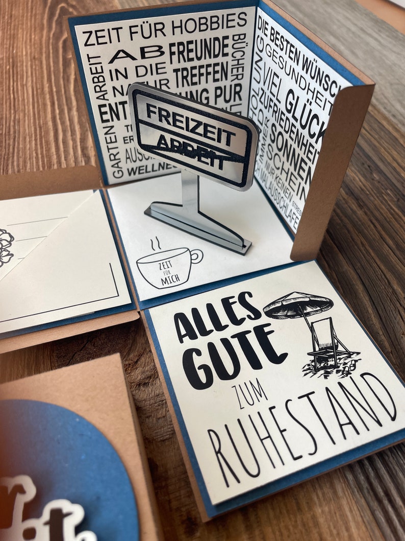 Puede incluir: Una tarjeta blanca con texto negro que dice "Alles Gute zum Ruhestand" con una ilustraci&oacute;n en blanco y negro de una sombrilla de playa y una silla de playa. La tarjeta es parte de una tarjeta emergente con un letrero plateado que dice "Freizeit Arbeit" y una peque&ntilde;a taza blanca con el texto "Zeit f&uuml;r mich".