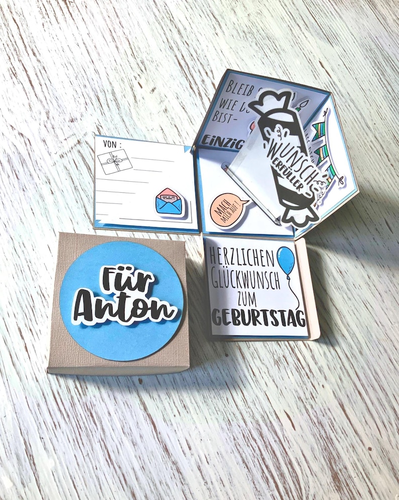 May include: A handmade birthday card with a blue and white color scheme. The card is in a box with the words "F&uuml;r Anton" on a blue circle. The card has multiple panels with German text and illustrations.