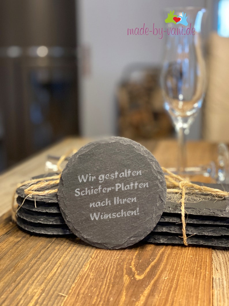 May include: A set of six round slate coasters with a natural, rough texture. The top coaster has the text "Wir gestalten Schiefer-Platten nach Ihren W&uuml;nschen!" engraved in white.