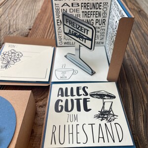Puede incluir: Una tarjeta emergente con fondo marr&oacute;n y detalles azules. La tarjeta presenta un cartel que dice "Freizeit Arbeit" y un mensaje que dice "Alles Gute zum Ruhestand".