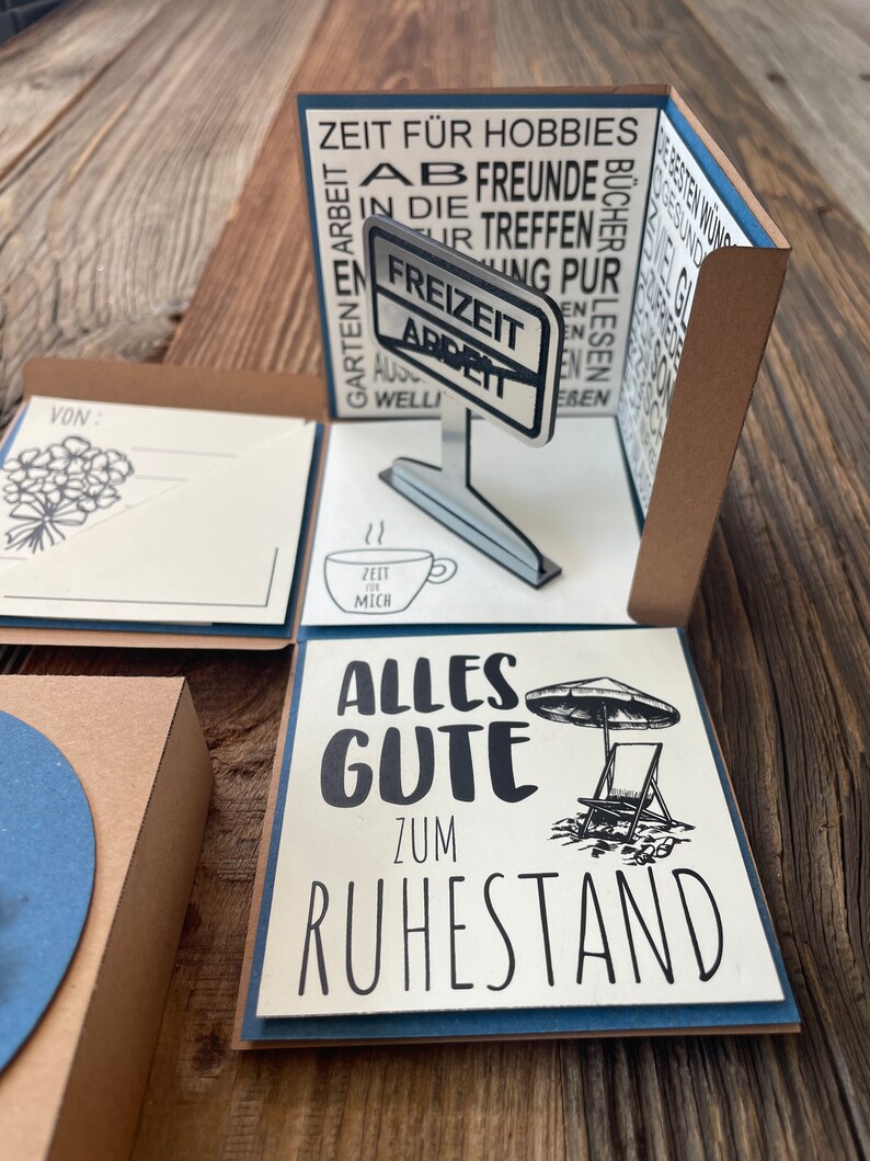 Puede incluir: Una tarjeta emergente con fondo marr&oacute;n y detalles azules. La tarjeta presenta un cartel que dice "Freizeit Arbeit" y un mensaje que dice "Alles Gute zum Ruhestand".