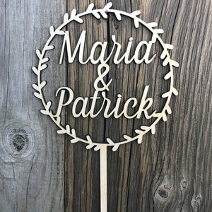 May include: A wooden cake topper with a circle of leaves and the text "Maria & Patrick" in a cursive font.