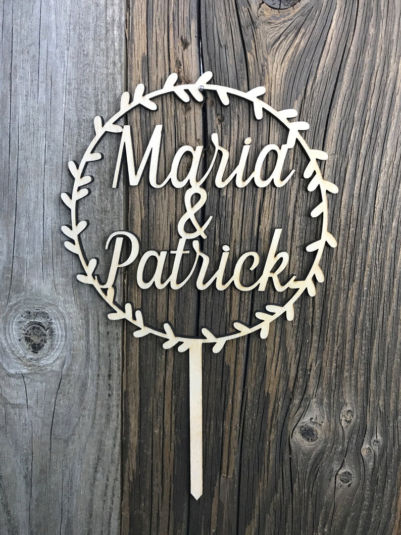 May include: A wooden cake topper with a circle of leaves and the text "Maria & Patrick" in a cursive font.