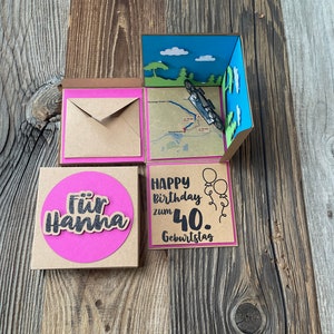 May include: A brown cardboard box with a pink and blue pop-up card inside. The card features a small toy car, a map, and the words "Happy birthday zum 40. Geburtstag". There is a separate brown card with a pink circle and the words "F&uuml;r Hanna".