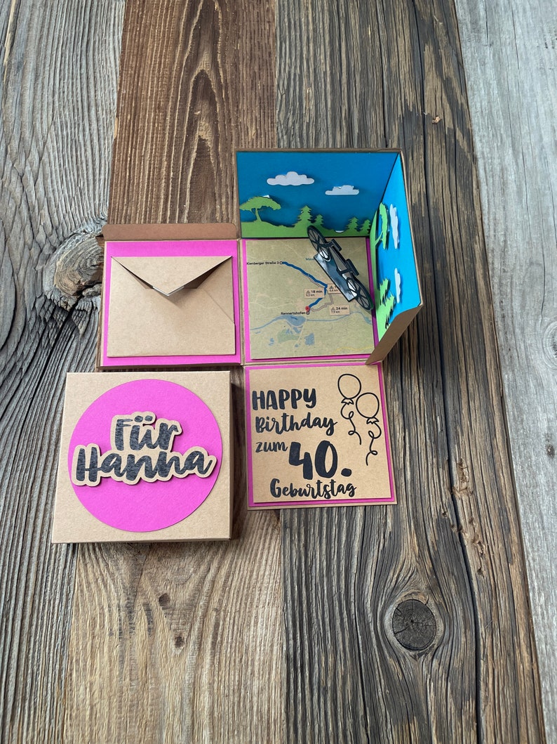 May include: A brown cardboard box with a pink and blue pop-up card inside. The card features a small toy car, a map, and the words "Happy birthday zum 40. Geburtstag". There is a separate brown card with a pink circle and the words "F&uuml;r Hanna".
