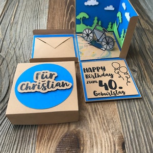 May include: A brown box with a blue circle on the front that says "F&uuml;r Christian". The box is open and there is a blue card inside that says "Happy Birthday zum 40. Geburtstag" with a bicycle and balloons.