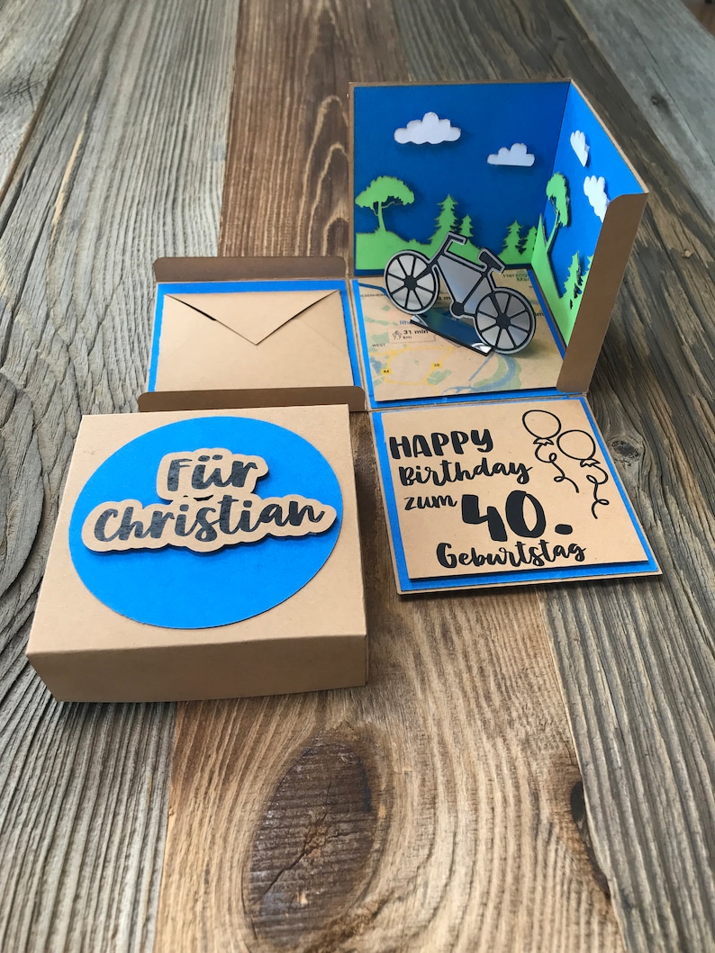May include: A brown box with a blue circle on the front that says "F&uuml;r Christian". The box is open and there is a blue card inside that says "Happy Birthday zum 40. Geburtstag" with a bicycle and balloons.
