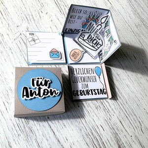 May include: A handmade birthday card with a pop-up design. The card is in a box with a blue circle that says "F&uuml;r Anton". The pop-up has birthday cake and balloon illustrations, with German text. The card is on a light wood surface.