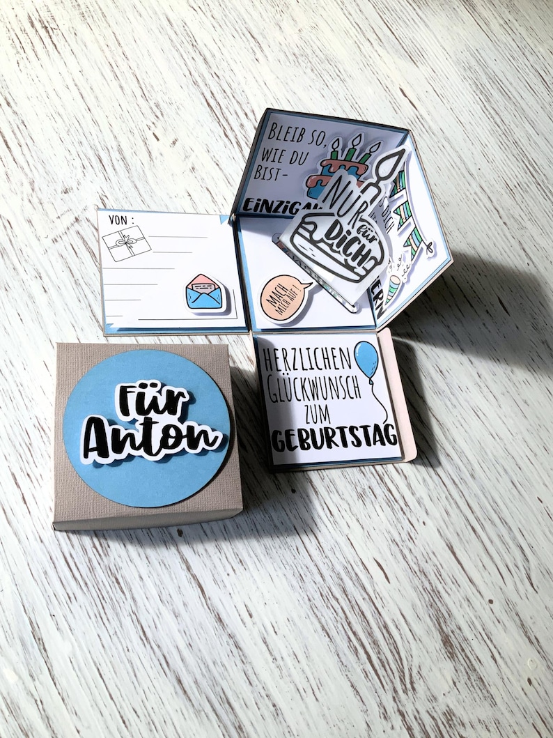 May include: A handmade birthday card with a pop-up design. The card is in a box with a blue circle that says "F&uuml;r Anton". The pop-up has birthday cake and balloon illustrations, with German text. The card is on a light wood surface.