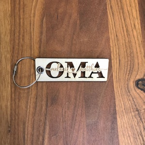 May include: Wooden keychain with the word "OMA" cut out and the names "Melanie" and "Matthias" written above and below the letters. The keychain has a silver metal ring.