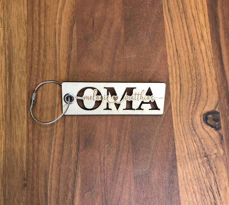May include: Wooden keychain with the word "OMA" cut out and the names "Melanie" and "Matthias" written above and below the letters. The keychain has a silver metal ring.