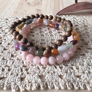 May include: Two stacked bracelets featuring a mix of round wooden beads with a dark brown grain pattern and various smooth, polished, multi-coloured gemstone beads in shades of pink, orange, purple, green, and translucent white.