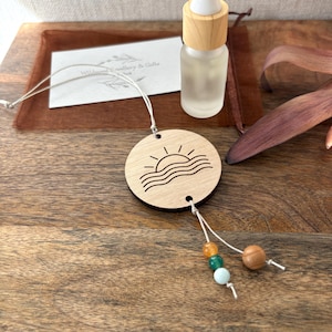 May include: A circular light wood pendant featuring a laser-engraved sun and wave design, suspended by a white cord. The cord is accented with one large wooden bead and three smaller beads in orange, green, and light blue, paired with a glass bottle.
