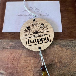 May include: A circular wooden ornament engraved with the words "CHOOSE happy" within a black rectangle. It features a sunflower design and hangs from a white cord with beads. A card with "Wildwood Jewelry & Gifts" is visible in the background.