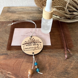 May include: A round wooden ornament engraved with a floral design and the text "YOU ARE enough," featuring a white cord with two blue beads and one wooden bead, accompanied by a small frosted glass bottle with a wooden dropper cap.