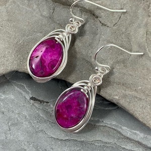 May include: A pair of silver wire wrapped earrings with pink agate stones. The stones have a swirling pattern and are set in a silver wire cage.