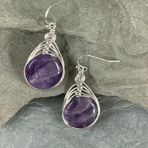 May include: A pair of silver wire wrapped earrings with amethyst stones. The stones are a deep purple color and have a natural, organic look. The earrings are a simple and elegant design.