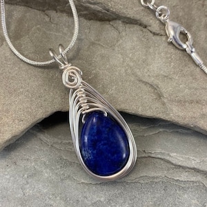 May include: A silver chain necklace with a teardrop-shaped pendant featuring a dark blue lapis lazuli stone wrapped in silver wire.